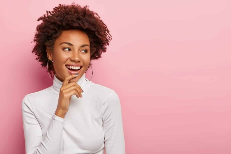 Satisfied carefree female model smiles gently, touches chin, looks aside, notices funny scene, laughs at something, has natural curly dark hair, dressed casually, isolated on pink background
